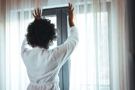 Beautiful african american woman in white bathrobe looking out the windowの写真素材