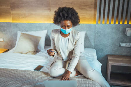Young african american businesswoman in medical mask using mobile phone while sitting on bed at hotel roomの写真素材