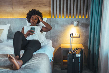 African american woman using mobile phone while sitting on bed in hotel roomの写真素材