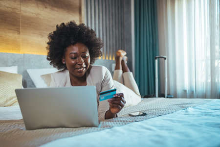 Smiling african american woman using laptop and credit card in hotel roomの写真素材