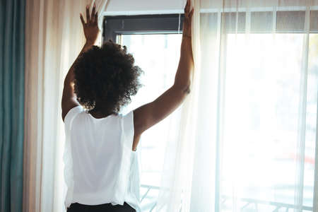Back view of young african american woman opening window at homeの写真素材