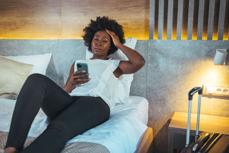 African american woman using mobile phone while sitting on bed in hotel roomの写真素材