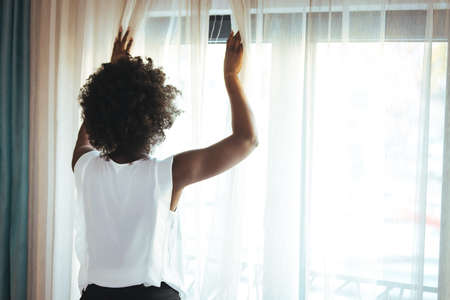 Young african american woman using mobile phone while standing near window at homeの写真素材