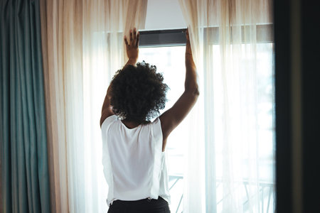 Back view of a young african american woman opening a window at home