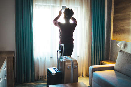 Rear view of young african american woman with suitcase and smartphone in hotel roomの写真素材