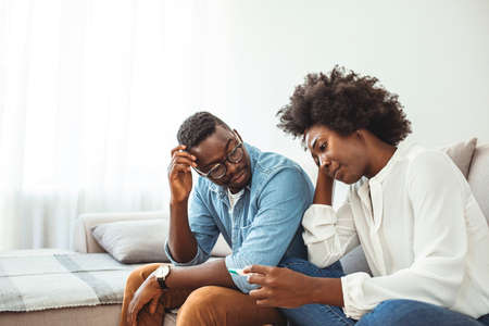 Sad african american couple looking at thermometer while sitting on sofa at homeの写真素材