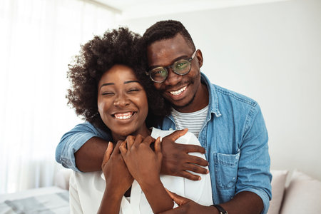 Cheerful black couple hugging and looking at camera while standing at home