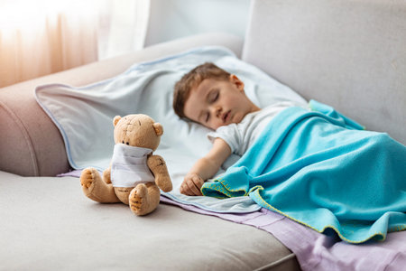 Sick little boy sleeping on sofa with teddy bear at home