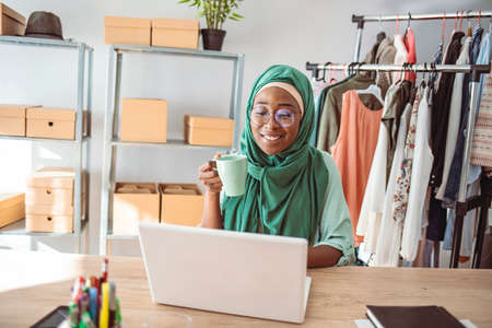 smiling african american woman in hijab holding cup of coffee and using laptop in officeの写真素材
