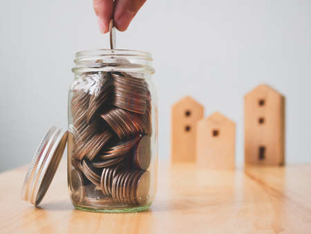 Property investment real estate and house mortgage financial concept. Hand putting money coins in jar with wooden home on tableの写真素材