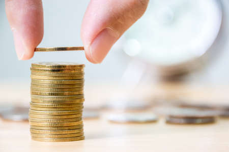 Hand putting gold coin stack with blurred clock background, Finance and investment save money conceptの写真素材