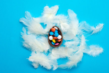 White feathers lie on the blue Easter background. Multi-colored candies in the chocolate egg