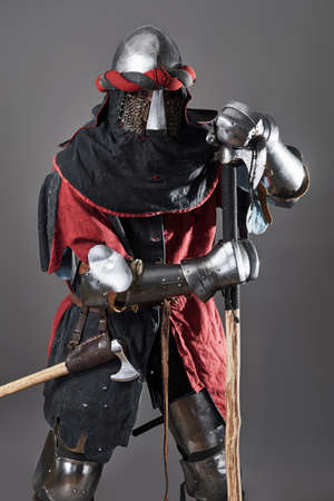 Medieval knight on grey background. Portrait of brutal dirty face warrior with chain mail armour red and black clothes and battle axeの写真素材