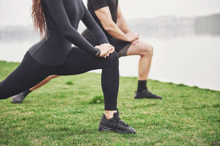 Fitness couple stretching outdoors in park near the water. Young bearded man and woman exercising together in morningの写真素材