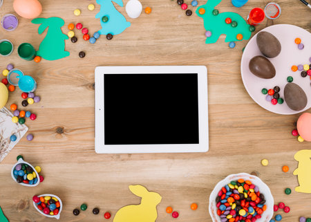 blank digital tablet surrounded with colorful gems candies easter eggs paper cutout bunny wooden...