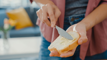 Hands of young Asian woman chef spreading butter on rustic rye bread with metal knife on wooden board on kitchen table in house. Fresh homemade bread production, Healthy eating and traditional bakery.の写真素材