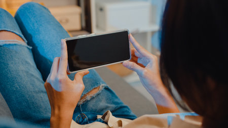 Young Asia lady use smart phone with blank black screen mock up display for advertising text while rest on couch in living room at modern home night. Chroma key technology, Marketing design concept.の写真素材