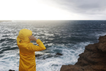 young woman dressed in yellow raincoat standing on the cliff looking on big waves of the sea while enjoying beautiful sea landscape in rainy day on the rock beach in cloudy spring weather.の写真素材