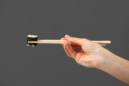 Woman hand holding fresh maki sushi roll with wooden chopsticks, isolated on grey background.の写真素材