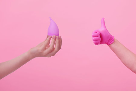 Hands of a young woman in pink latex gloves and hand with menstrual cup on pink background. women's health. preservation of ecology. waste-free use. women's hygiene products.の写真素材
