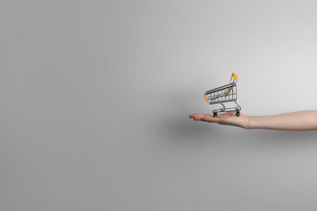 closeup of a woman's hand holding a small metal empty shopping cart in the palm of her hand on gray...
