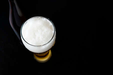 A glass of cold beer with foam and a bottle in the background. Black dark background with copy space.の写真素材