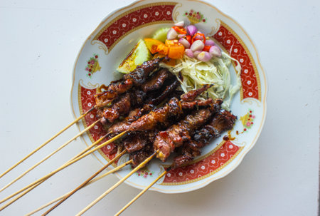 sate kambing or sate klatak or lamb satay or goat meat satay served with served with sliced ââcabbage, ââred onion, ââchili, ââtomatoes with soy sauce on plate. isolated white background -の素材