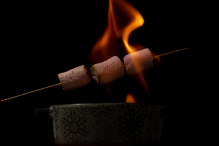 skewer of sweet marshmallows warming in the fire on a black backgroundの素材