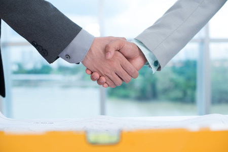 Male workers shaking hands over industrial blueprints and level