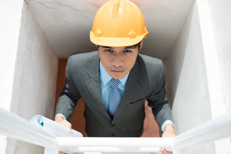 Serious supervisor climbing the ladder holding rolled blueprintsの写真素材