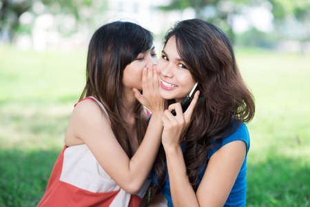 Image of a teenage girl sharing secret with her friend who talks over the phoneの写真素材