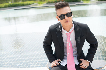 Young white-collar worker in sunglasses sitting on skateboard and listening to music during the...