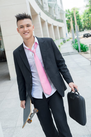 Vertical portrait of a smiling office worker walking along the street carrying skateboard and briefcaseの写真素材