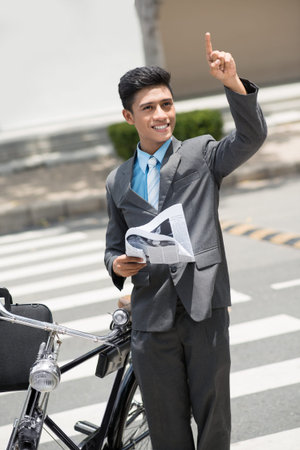 Vertical image of a young businessman standing with a newspaper and a bicycle outside and showing at...