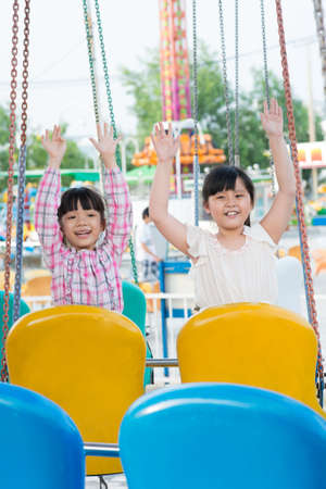 Vertical image of female youngsters sitting on swings with their hands raisedの写真素材