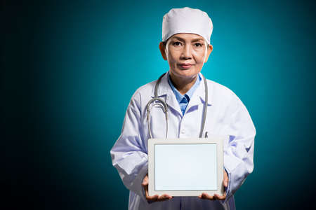 Copy-spaced portrait of a mature female doctor holding a tablet with empty screen isolated on blueの写真素材