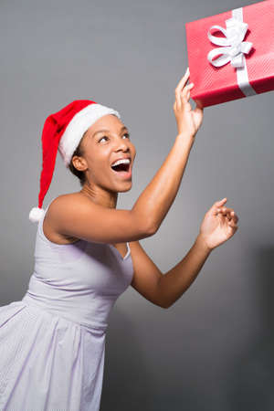 Vertical image of an excited lady in Santa hat with a x-mas present in hands over a grey backgroundの写真素材