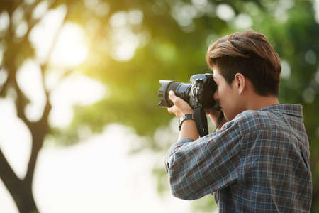 Handsome young man taking a photograph with his digital camera while standing outdoorsの写真素材