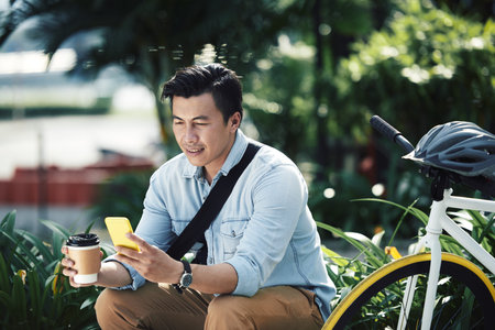 Asian man drinking take-out coffee and checking phone when sitting outdoors