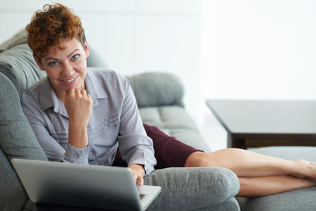 Pretty cheerful business woman lying on sofa and working on laptopの写真素材