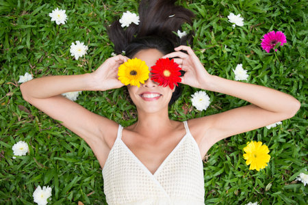 Angle view of a woman holding gerbera flowers on her eyes while lying on the grassの写真素材