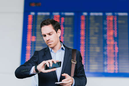 Serious businessman standing near airline schedule and looking at his watchの写真素材