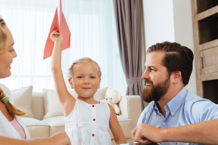Happy girl with a red paper airplane and her parents at homeの写真素材