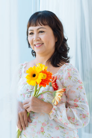 Smiling woman with flowers looking out of the windowの写真素材