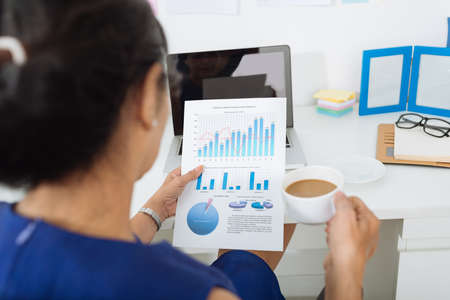 Business woman drinking coffee and examining financial document, view from the shoulderの写真素材