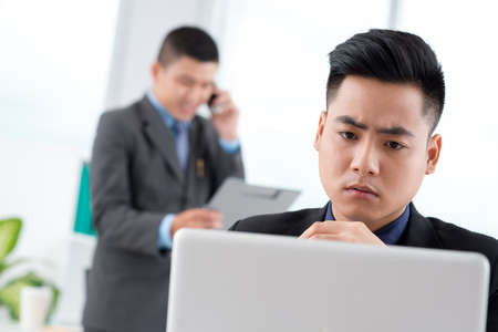 Serious Vietnamese manager reading something on the laptop, while his colleague talking on cellphone in backgroundの写真素材