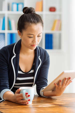 Serious businesswoman with a cup of tea reading something on digital tabletの写真素材