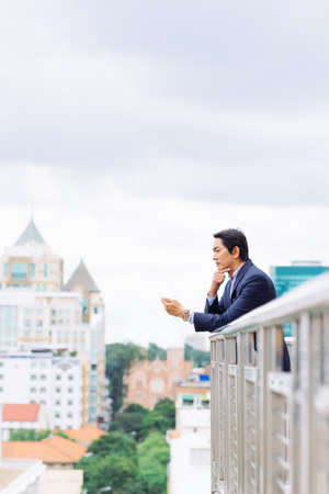 Pensive Asian businessman with a phone looking at the city from the roofの写真素材