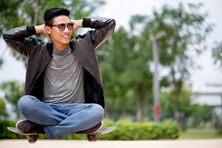 Young man sitting on the skateboard with hands behind his headの写真素材