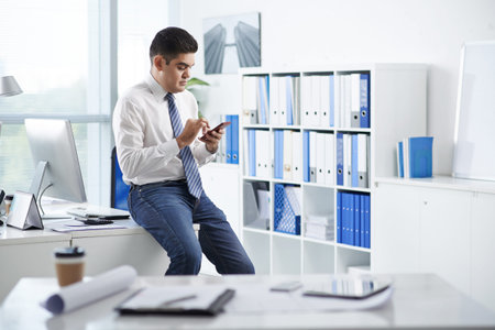 Businessman sitting on the edge of his desk and text messaging